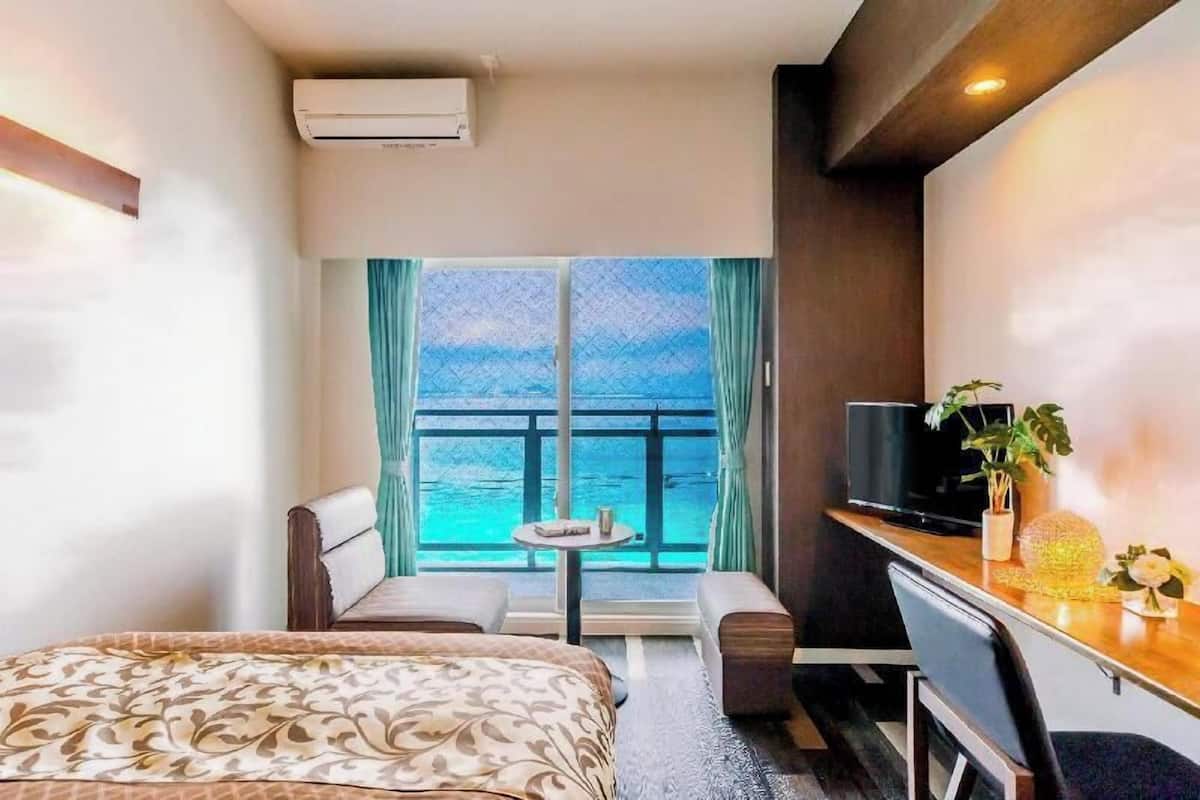 A hotel room with a bed, a desk, a TV, a small table, and a view of the sea through large windows.