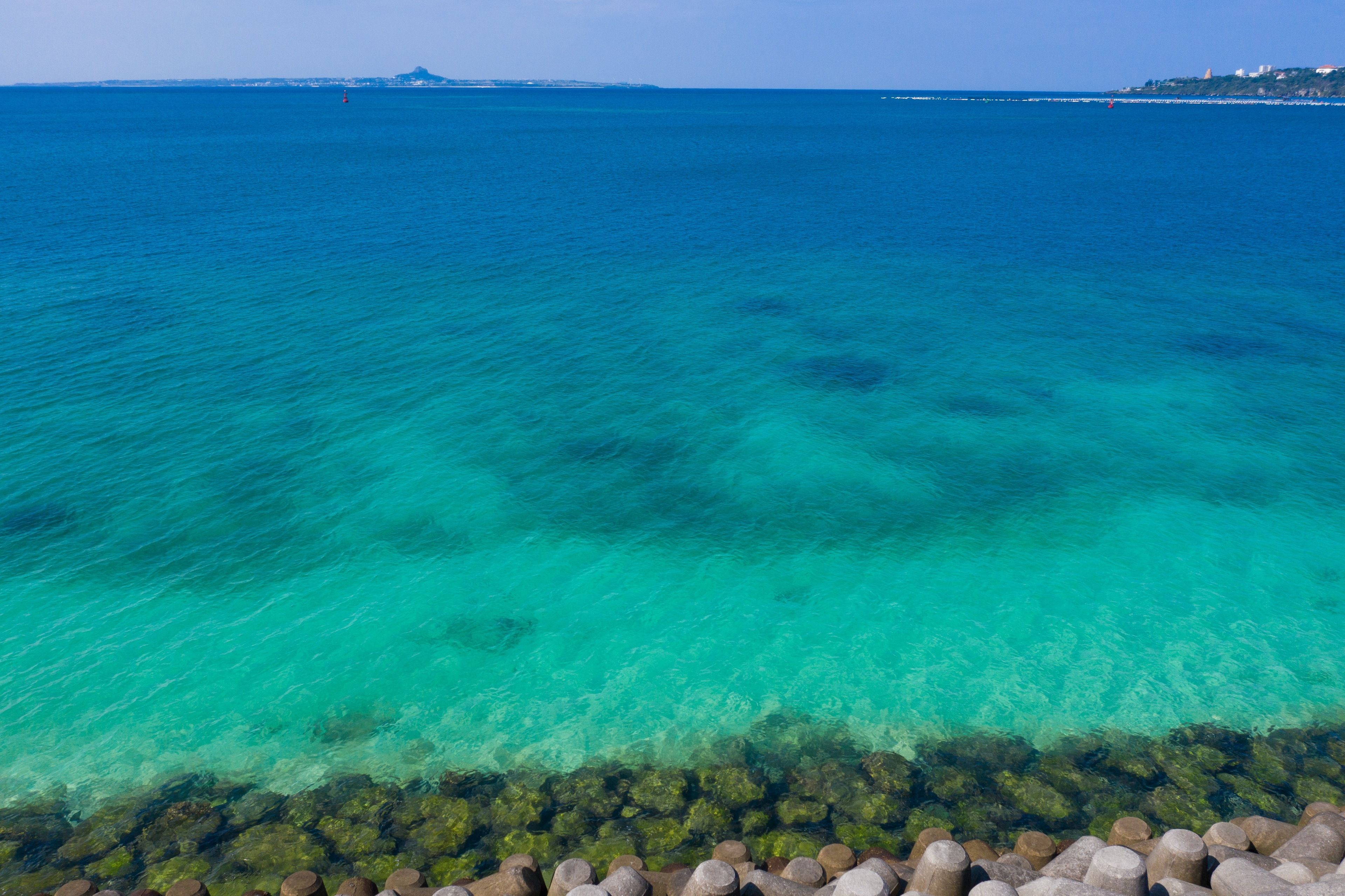 Vista costera con agua turquesa cristalina, un litoral rocoso y edificios a lo lejos.