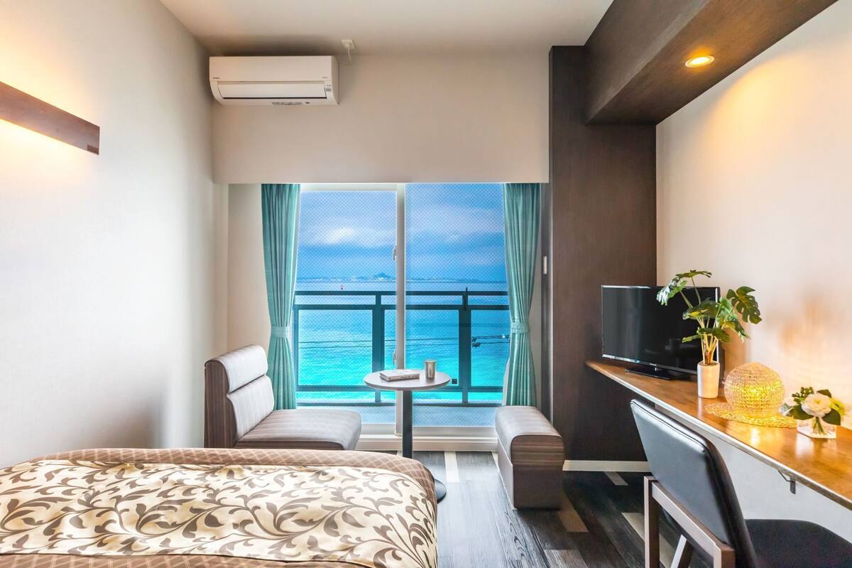 A hotel room with a bed, a desk, a TV, a small table, and a view of the ocean.