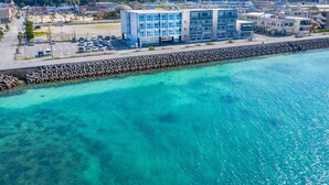 An aerial view of a coastal area with a clear blue sea, a rocky breakwater, and a modern building with multiple floors and large windows. - Mr.KINJO in MOTOBU OCEAN TERRACE (Motobu)