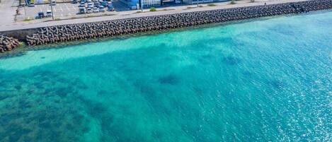 A coastal hotel with a clear view of the turquoise sea, a parking lot, and a modern building with multiple floors.