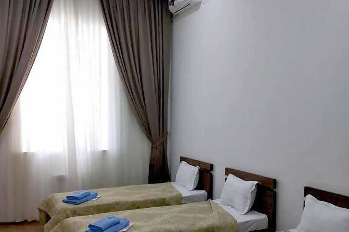 Une chambre d’hôtel avec trois lits simples, chacun équipé d’une serviette bleue, d’oreillers blancs et d’une tête de lit en bois. Un climatiseur est fixé au mur, et la chambre possède un plancher en bois et une fenêtre avec des rideaux.