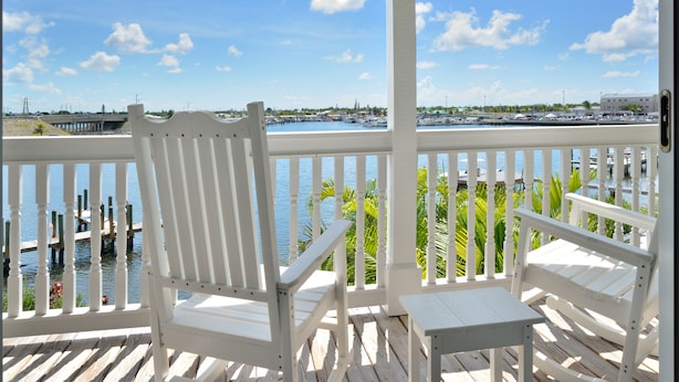 A wooden deck with white chairs and a table, overlooking a marina with boats and a bridge in the background. - Luxury 3-Bedroom Waterfront Home with Pool AND Dock! (Key West)