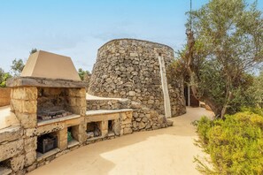 A stone structure with a flat-topped table and a stone bench, surrounded by greenery and a clear sky.