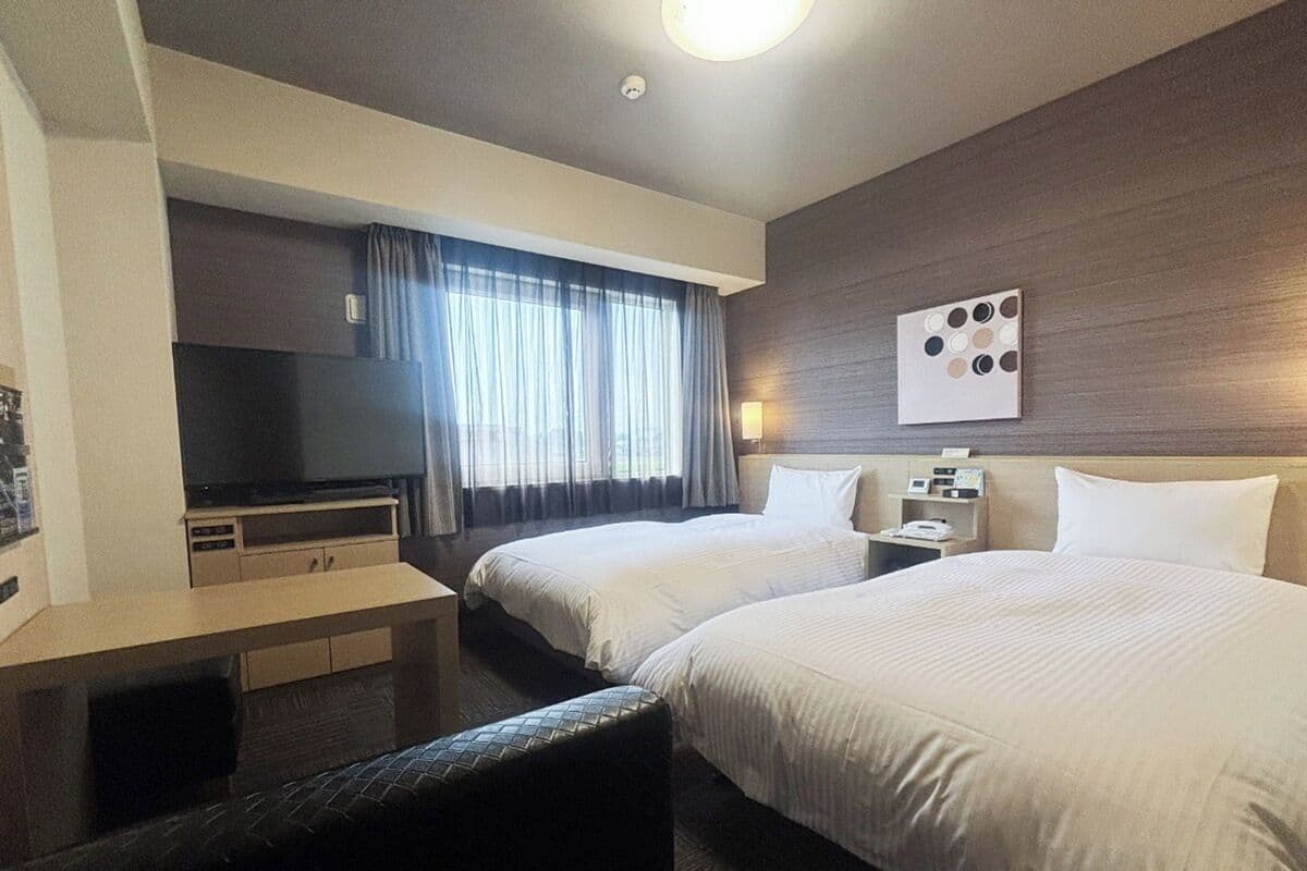 Ein Hotelzimmer mit zwei Betten, einem Schreibtisch, einem Fernseher und einem Fenster mit Vorhängen.