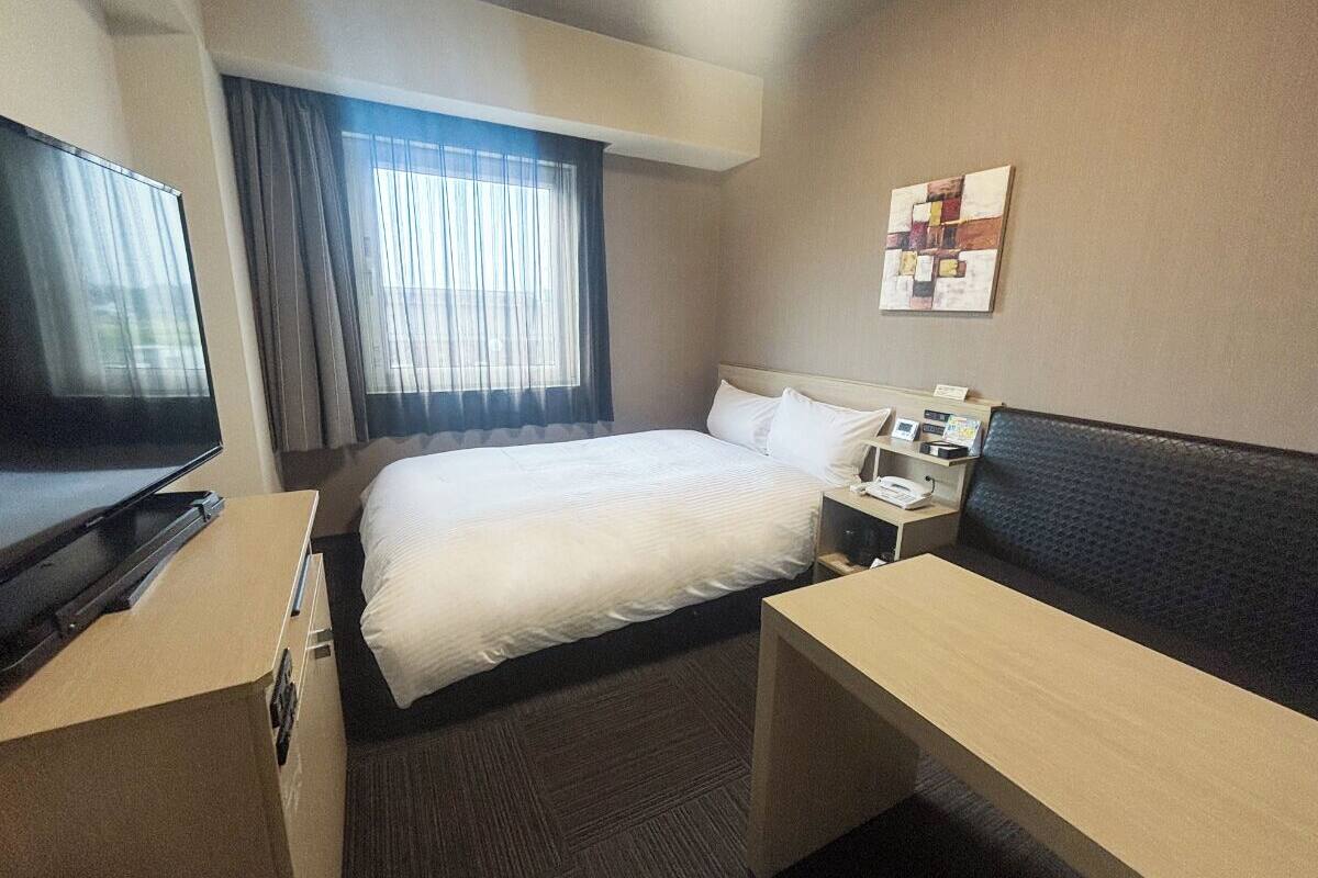 Ein Hotelzimmer mit Bett, Schreibtisch, Fernseher, einem Fenster mit Vorhängen und einem an der Wand hängenden Kunstwerk.