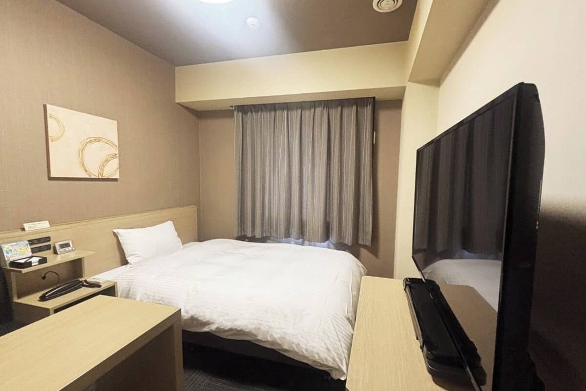 Ein Hotelzimmer mit einem Bett, einem Schreibtisch mit Telefon, einem Fernseher und einem Fenster mit Vorhängen.