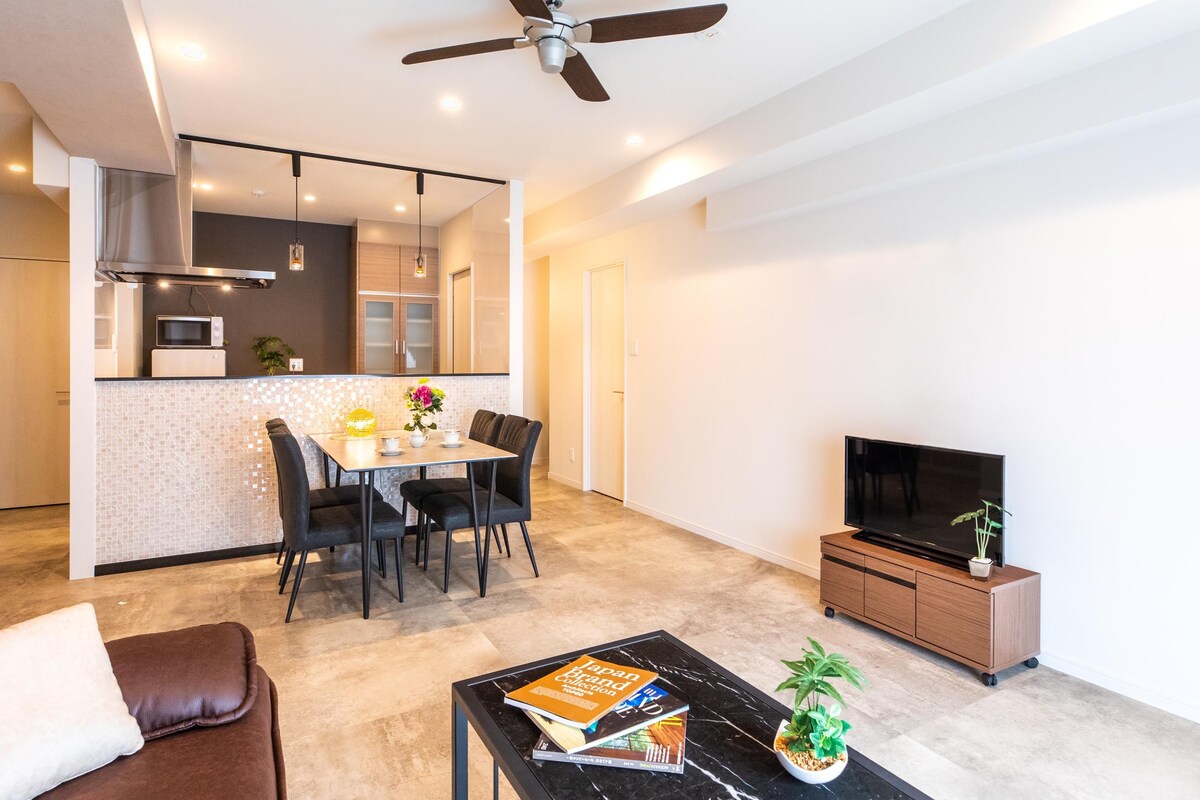 A modern living room with a flat-screen TV, a coffee table with books and a plant, a dining area with a table and chairs, and a kitchen with a microwave and stove.