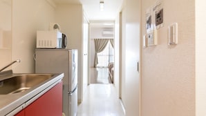 A narrow hallway with a kitchen area on the left, featuring a sink, a microwave, and a refrigerator. There is a door leading to a bedroom with a bed and curtains. - Mr.KINJO PREMIUMHOME in CHATAN 2 (Chatan)