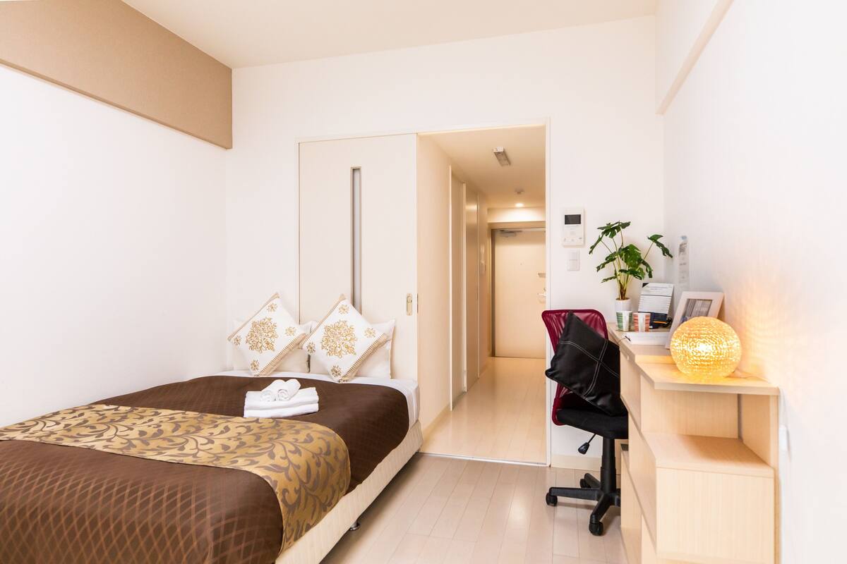 A hotel room with a bed, a desk with a lamp, a chair, and a plant.