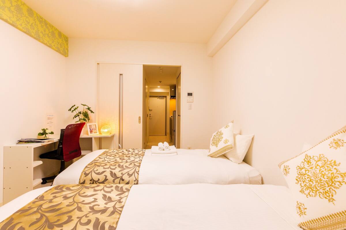 A hotel room with two beds, a desk, a chair, a plant, and a door leading to another room.