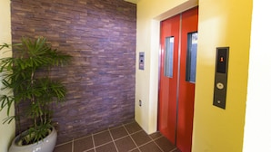 A hallway with a stone wall, a potted plant, a red elevator door, and a wall-mounted elevator control panel. - Mr.KINJO PREMIUMHOME in CHATAN 2 (Chatan)