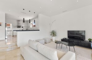 A modern living room with a sofa, a coffee table, and a TV. There is a kitchen in the background with a dining area. - Villa near Beach with Pool & Panoramic Views (Marbella)