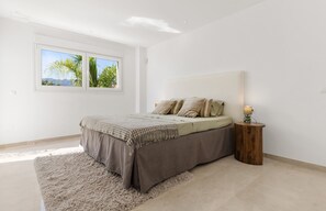 A bedroom with a bed, a nightstand, a lamp, and a view of palm trees through the window. - Villa near Beach with Pool & Panoramic Views (Marbella)