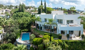 A modern two-story house with a swimming pool, surrounded by lush greenery and a hilly landscape.