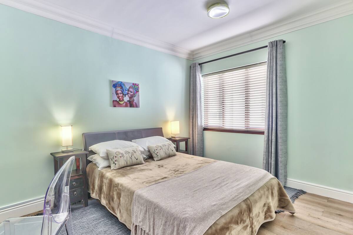 A bedroom with a bed, two bedside tables with lamps, a window with curtains, and a wall-mounted picture.
