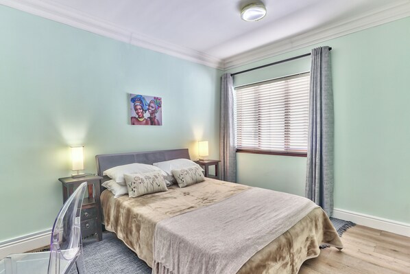 A bedroom with a bed, two bedside tables with lamps, a window with curtains, and a wall-mounted picture. - Planet Africa (Cape Town)