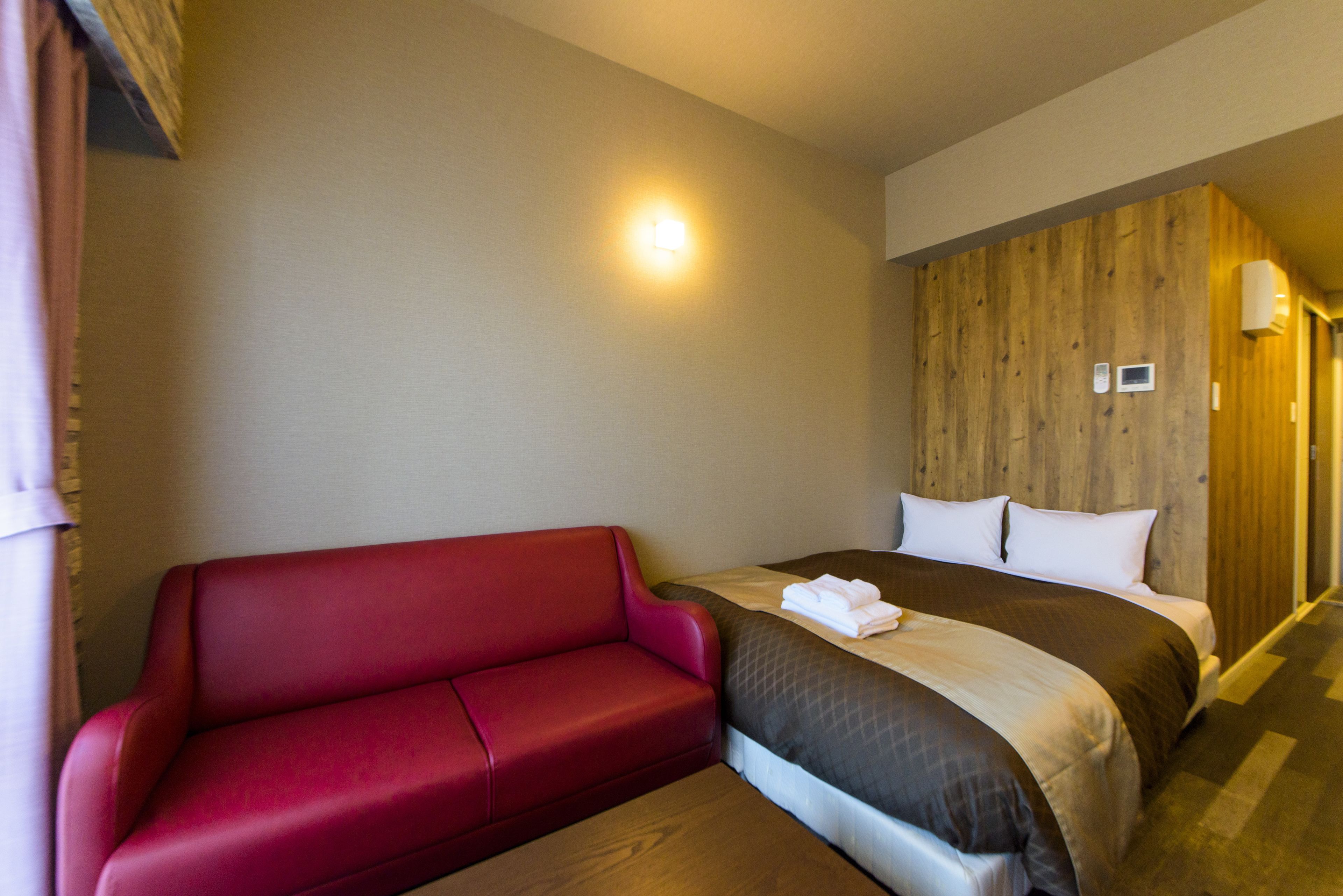 Habitación de hotel con una pared de acento de madera, un sofá rojo, una cama con edredón marrón y blanco, y una mesita de noche con lámpara.