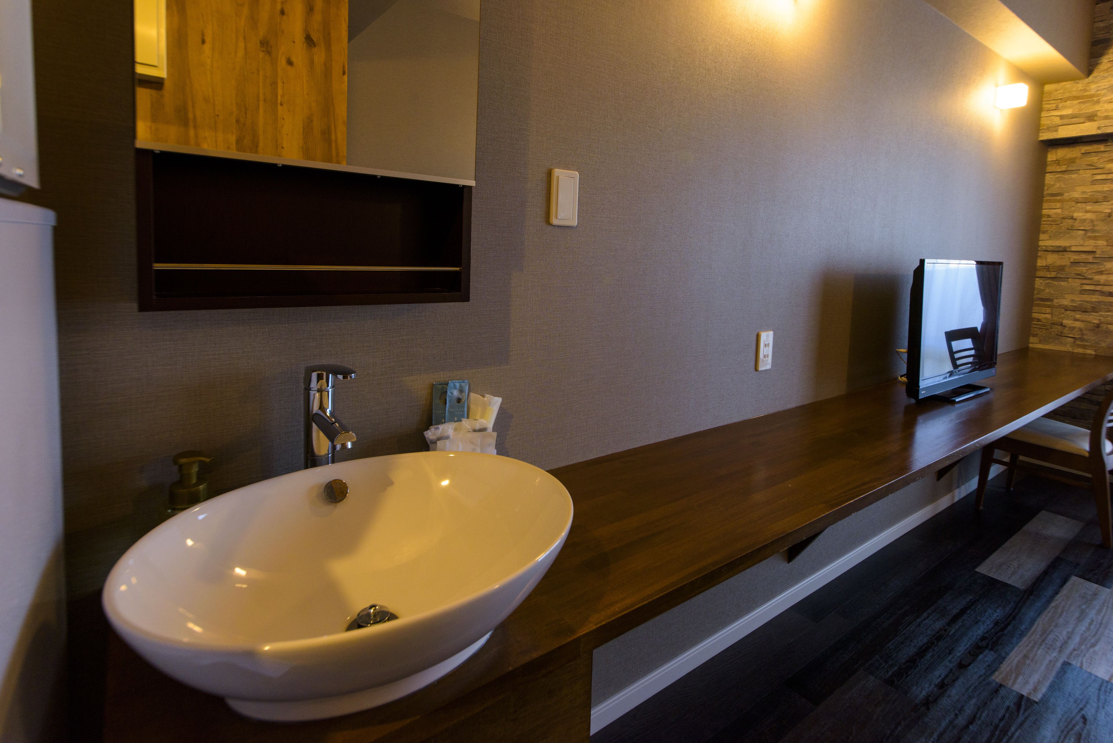 Un baño con un lavamanos grande y blanco, encimera de madera y un espejo encima. Hay un estante pequeño sobre el espejo y un televisor a la derecha.