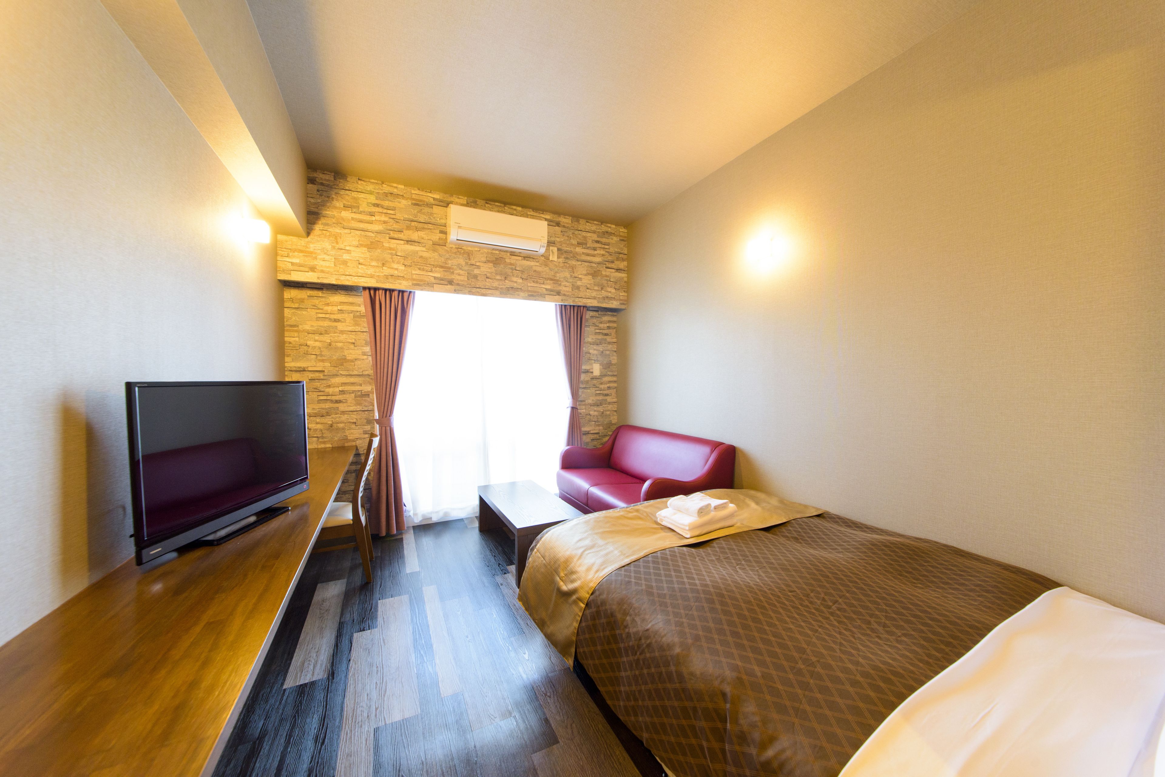 A hotel room with a bed, a TV, a red sofa, and a stone wall.