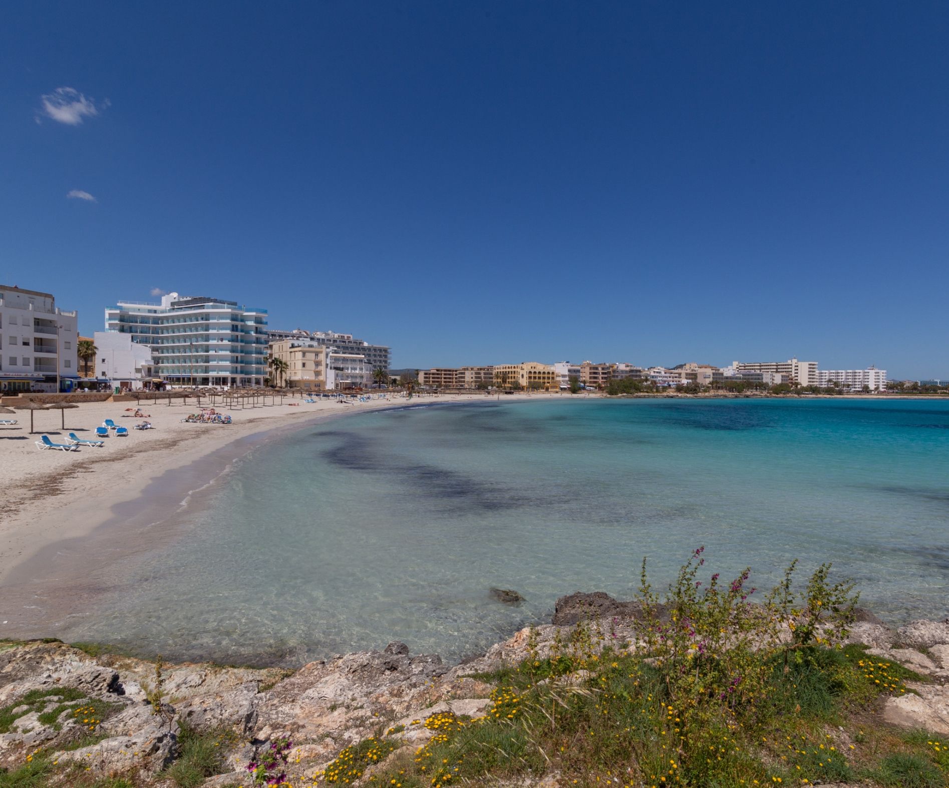A beach with clear turquoise waters, a sandy shore, and a row of buildings with balconies overlooking the sea.