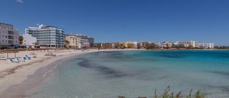 A beach with clear turquoise waters, a sandy shore, and a row of buildings with balconies overlooking the sea.