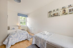 Un dormitorio con dos camas, una ventana con persianas azules, un radiador, una mesita de noche con una lámpara y un cuadro en la pared.