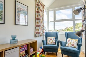 A room with two blue armchairs, a wooden desk with children's toys, a framed picture, and a view of a town through a large window.