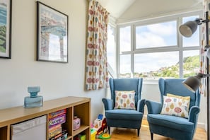 Una habitación con dos sillones azules, un escritorio de madera con juguetes infantiles, un cuadro enmarcado y vistas a un pueblo a través de ventanas amplias.