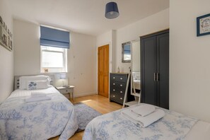 A bedroom with two beds, a dresser, a wardrobe, a window with blinds, and a bedside table with a lamp.