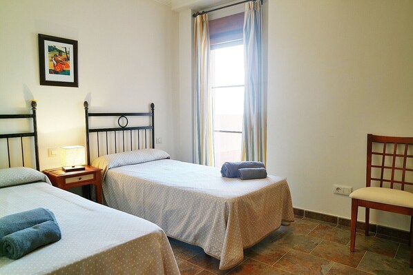 A room with two beds, a chair, a nightstand with a lamp, and a window with curtains.