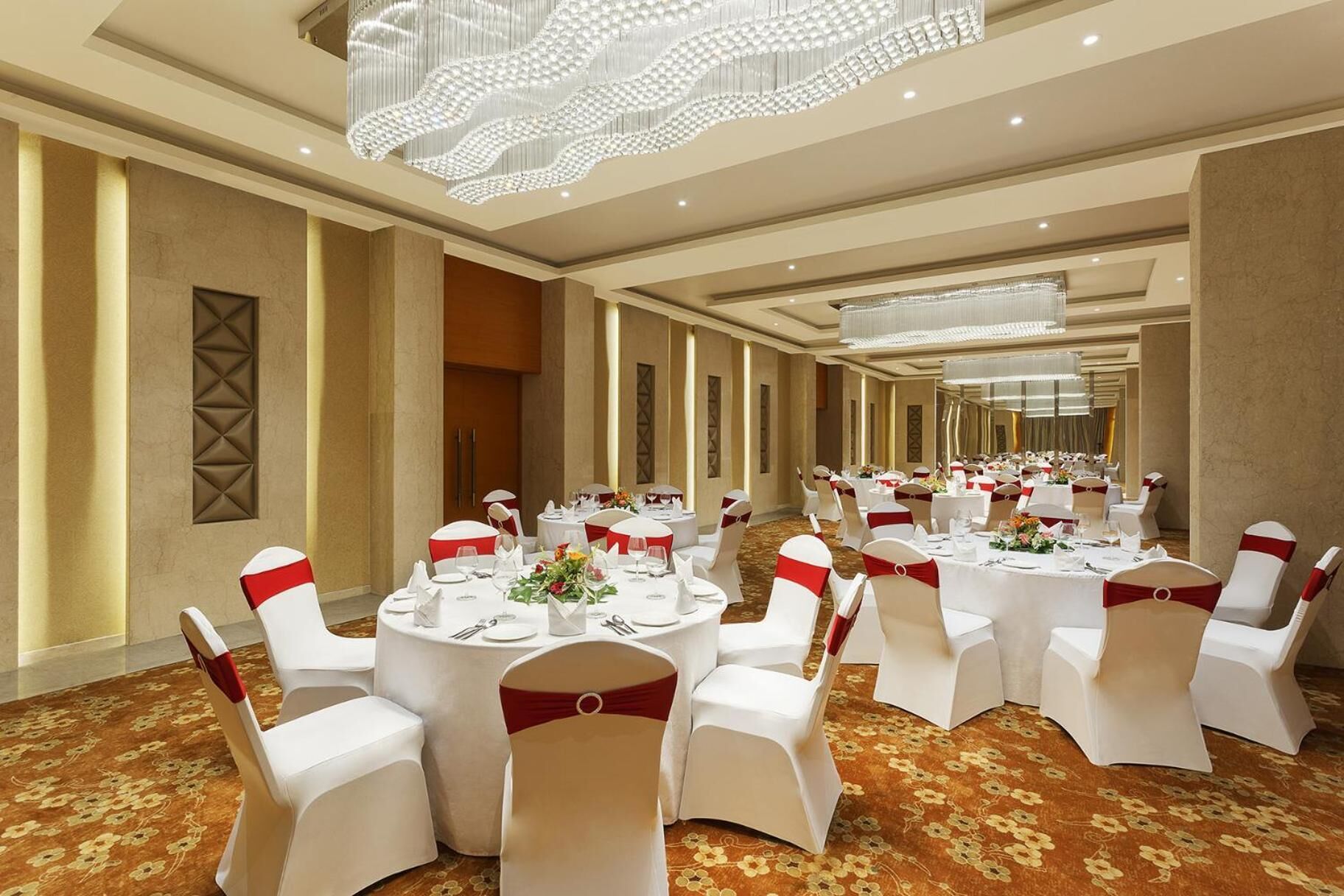 A banquet hall with round tables, white chairs with red sashes, and a large chandelier.