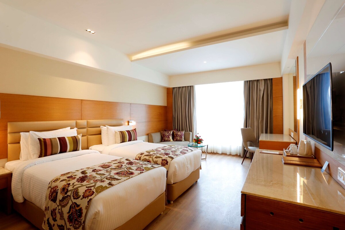 A hotel room with two beds, a desk, a TV, and a window with curtains.