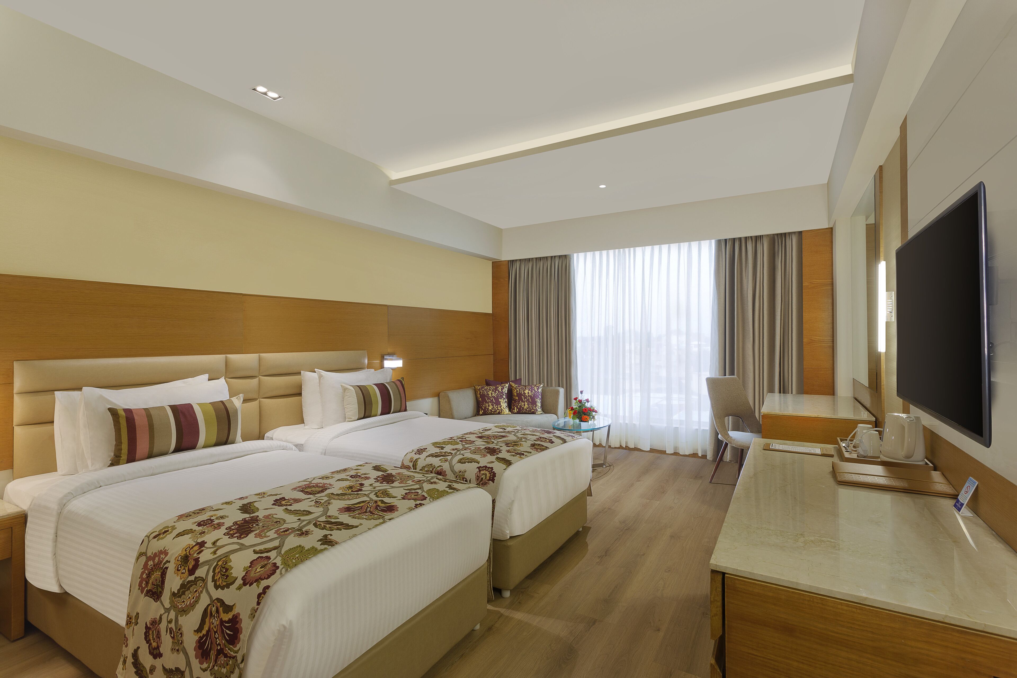 A hotel room with two beds, a desk, a TV, and a large window with curtains.
