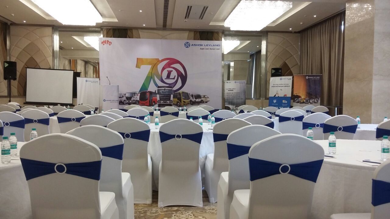 A conference room with rows of white chairs with blue sashes, a large banner with a logo and text, and a projector screen.
