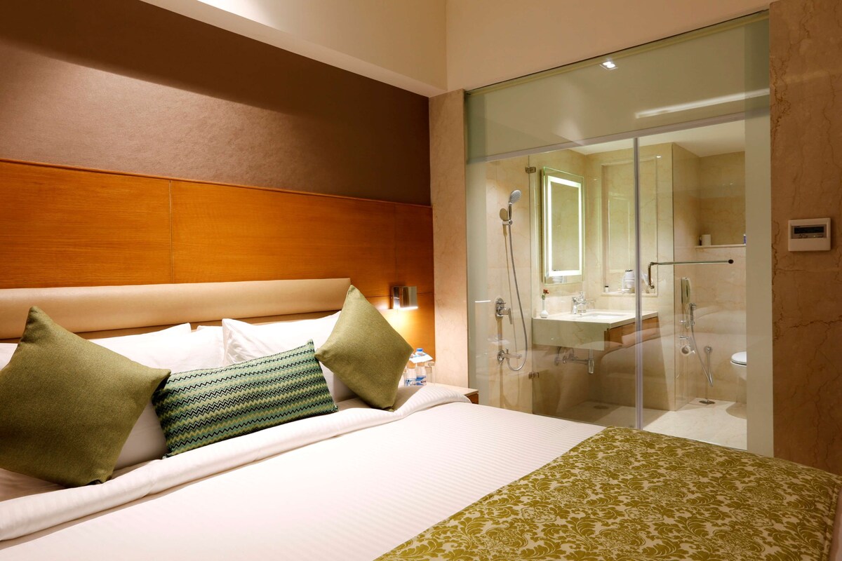A hotel room with a bed, pillows, a bedside table, a glass-enclosed shower area, and a bathroom with a sink and mirror.