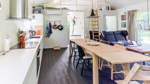A compact, modern kitchen with a wooden dining table, black chairs, and a central island. A lofted sleeping area with a ladder is visible in the background. - 3 bedroom awesome home in Højby (Højby)