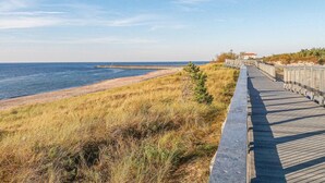 A wooden boardwalk leading to a beach with a distant pier and a building with a red roof.
