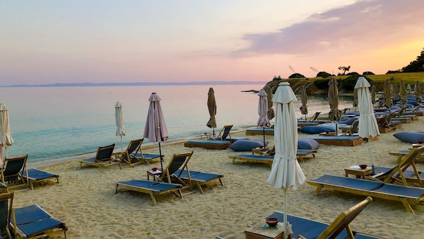 A beach with rows of sun loungers and umbrellas, a calm sea, and a distant shoreline. - Alexanika Apartments (Sithonia)