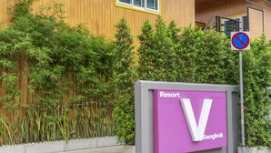 Une enseigne indiquant « Resort Bangkok », avec un grand logo en forme de « V », un panneau interdisant le stationnement, et un bâtiment en bois avec un cadre de fenêtre jaune en arrière-plan.