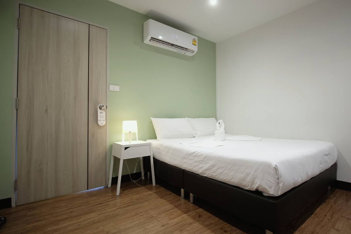 A hotel room with a bed, bedside table, nightstand, and an air conditioning unit.