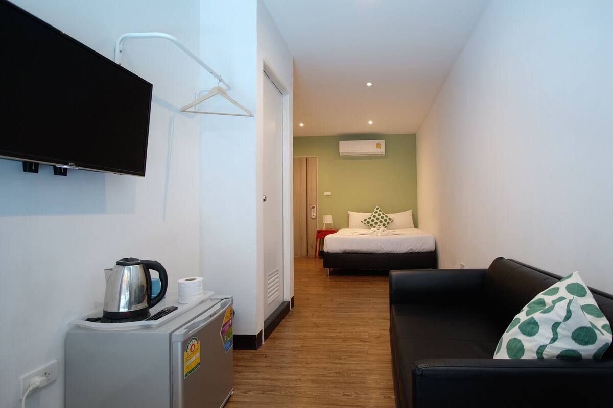 A narrow hallway leading to a bedroom with a bed, a black sofa with a green pillow, a small table with a kettle, and a TV mounted on the wall.