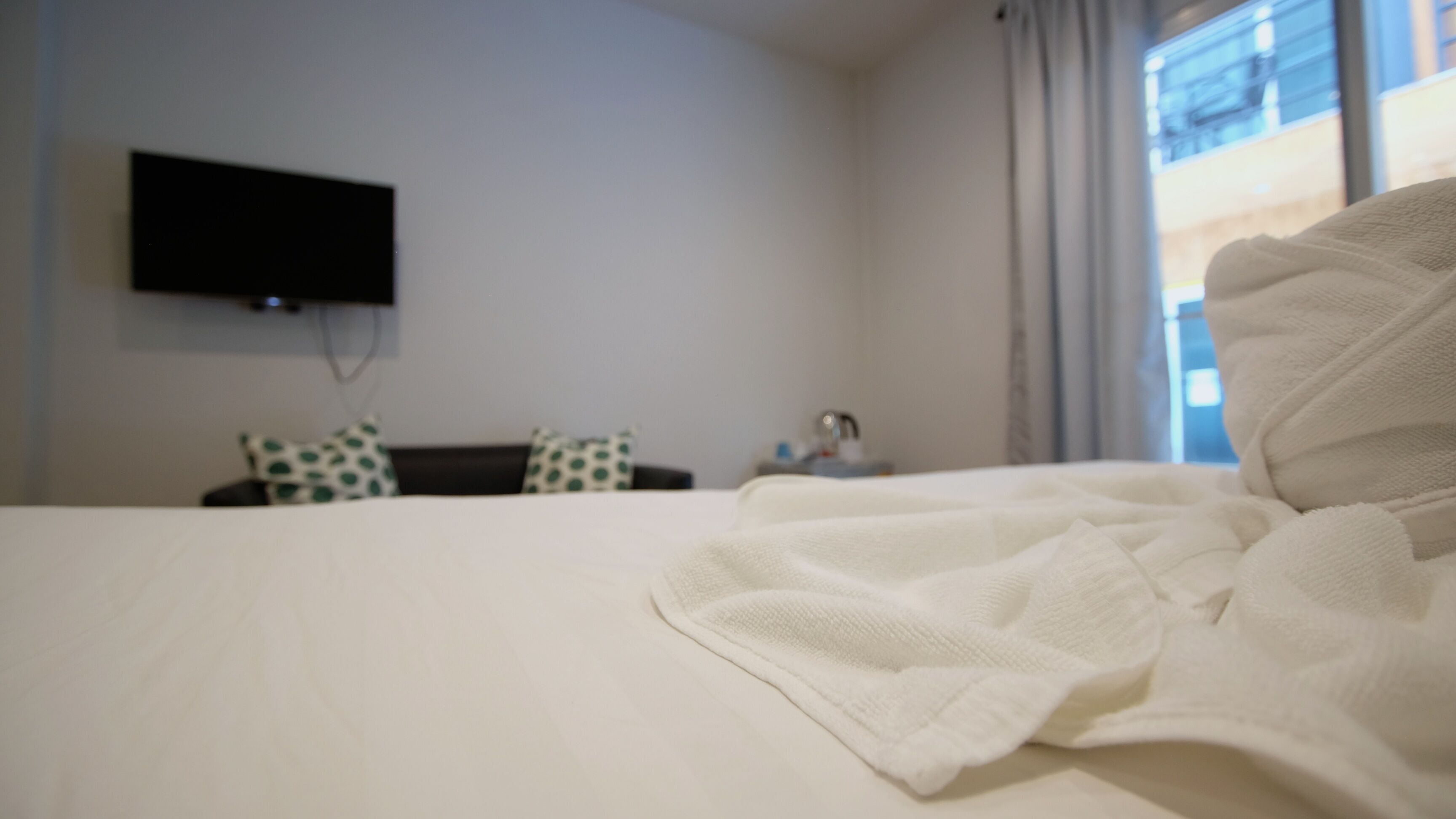 A hotel room with a bed, a flat-screen TV mounted on the wall, a nightstand, and a window with curtains.