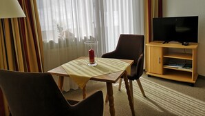 Ein Hotelzimmer mit einem Holztisch, zwei Stühlen, einem Fernseher auf einem Holzschrank und einem Fenster mit durchscheinenden Vorhängen.