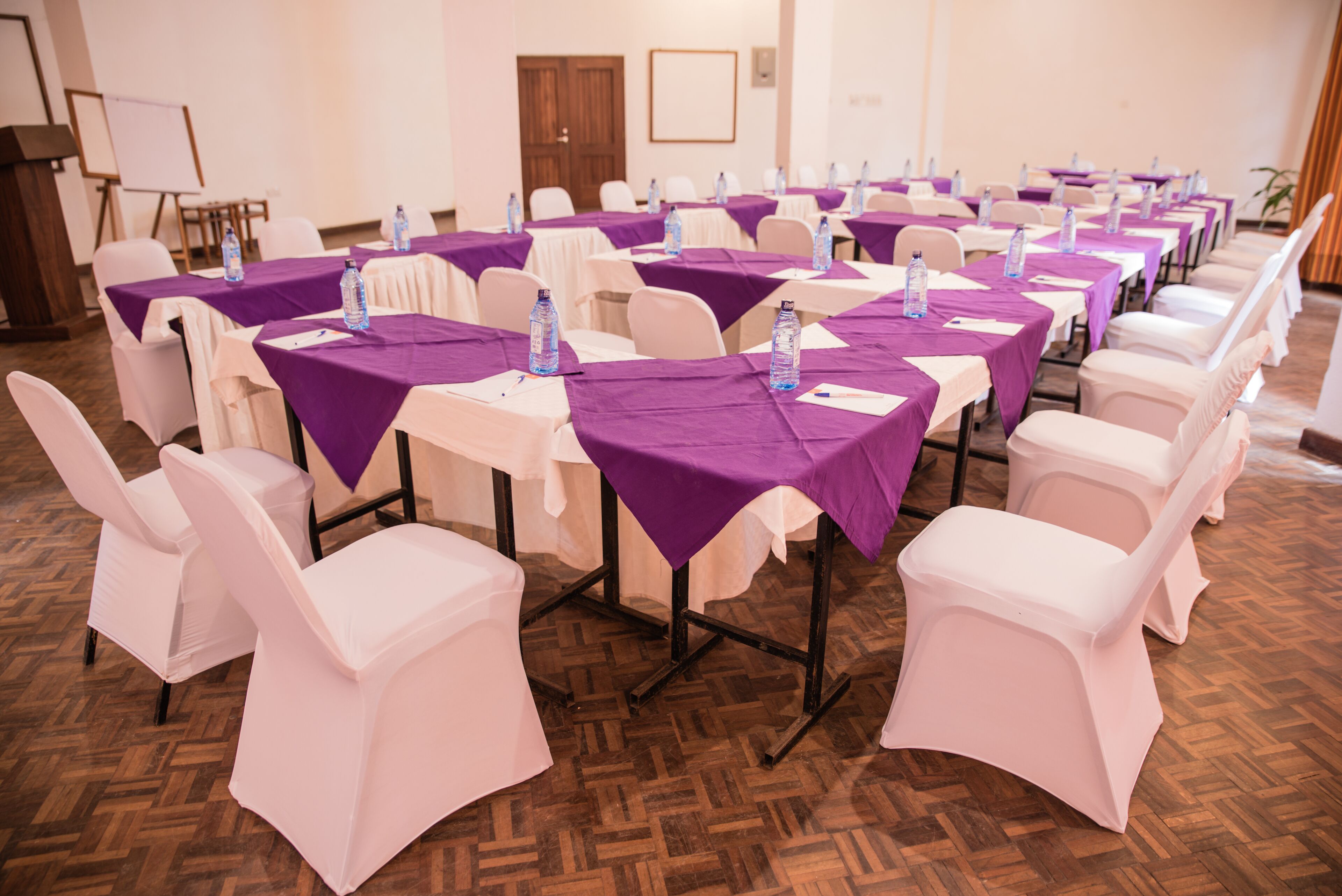 Una sala conferenze con diversi tavoli, ognuno ricoperto da una tovaglia viola e dotato di bottiglie d'acqua.