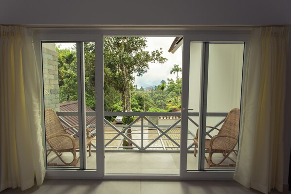 A room with a large sliding glass door leading to a balcony with wicker chairs and a view of lush greenery and a distant mountain.