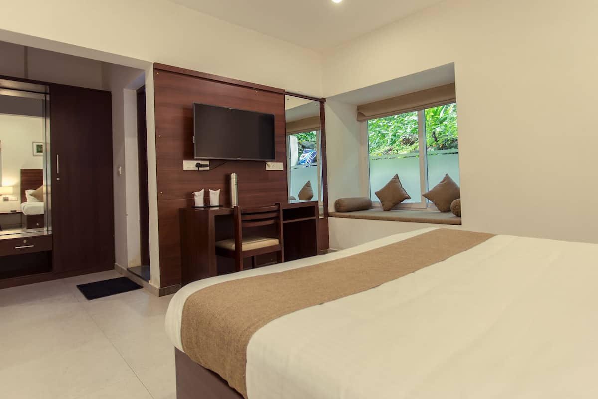 Habitación de hotel moderna con una cama grande, un televisor montado en un panel de madera, una zona de estar con cojines y vistas a zonas verdes a través de la ventana.