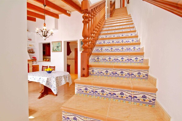 A staircase with ornate wooden balusters and patterned tiles, leading to a dining area with a table set for two and a chandelier.