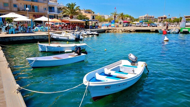 A marina with several small boats docked, a wooden pier, and buildings with outdoor seating in the background.