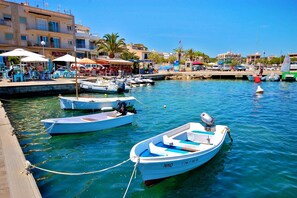Un port de plaisance où sont amarrés plusieurs petits bateaux, une jetée en bois et, au fond, des bâtiments dotés de terrasses extérieures.
